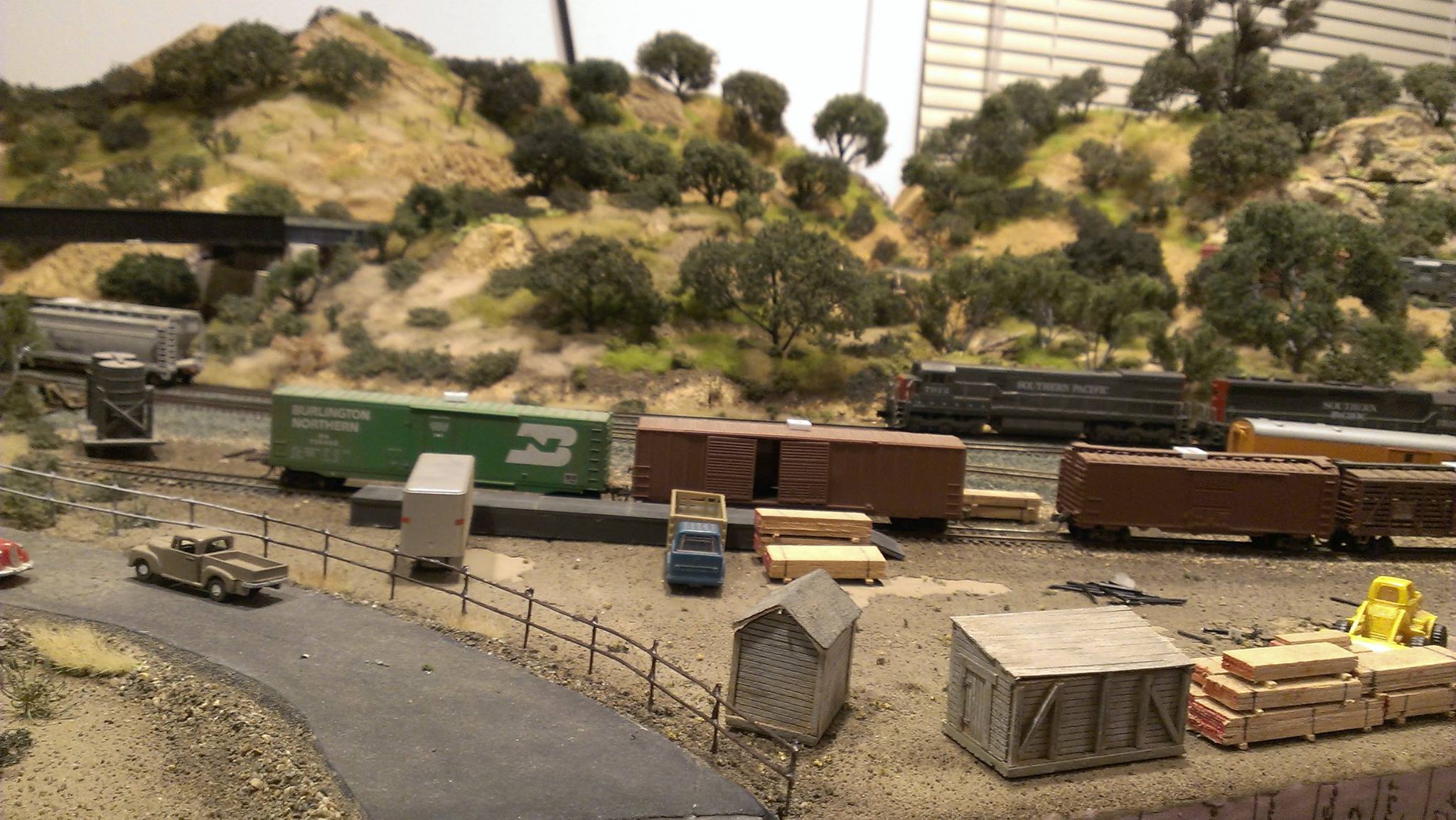THE TEAM TRACK AREA ON MY N SCALE MODEL RAILROAD. SOME BOX CARS ARE SITTING AT THE UNLOADING RAMP, AND BEHIND THEM A PAIR OF SOUTHERN PACIFIC DIESELS ARE PULLING A TRAIN UPGRADE. IN THE DISTANCE, THE ROLLING HILLS OF CALIFORNIA ARE UNMISTAKEABLE WITH THE BLUE OAKS DOTTING THE LANDSCAPE,